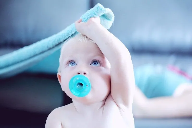 Baby with a dummy in its mouth pulling a blanket over its head.