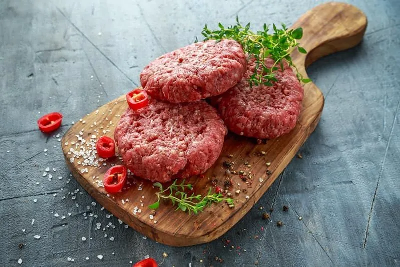 Raw hamburger patties on a chopping board with herbs sprinkled on top.