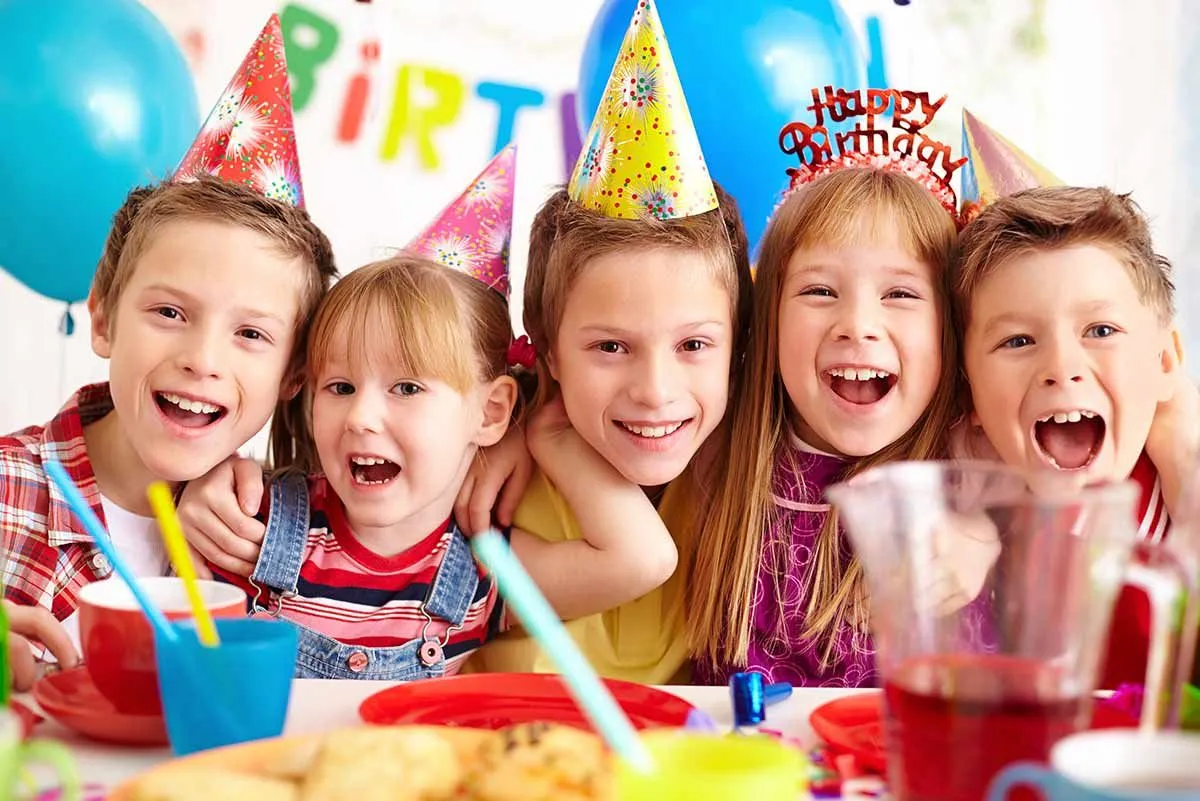 Five kids at a birthday party wearing party hats, smiling and excited for the airplane cake.