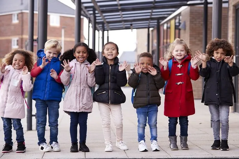 Group of toddlers standing and smiling in a row wearing coat and schoolbag.