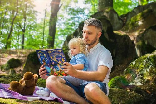 A man is sat outdoors in a shaded woodland area, reading to his son on his lap.